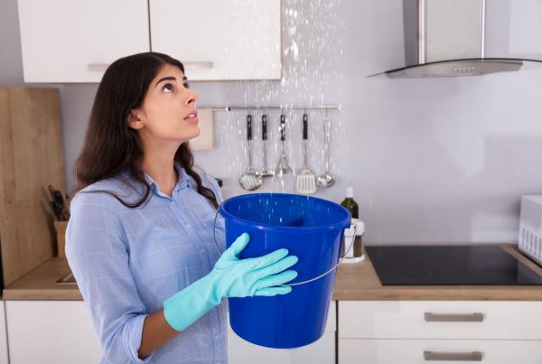 Worried Woman Holding Bucket While Water Droplets Leak From Ceiling In Kitchen